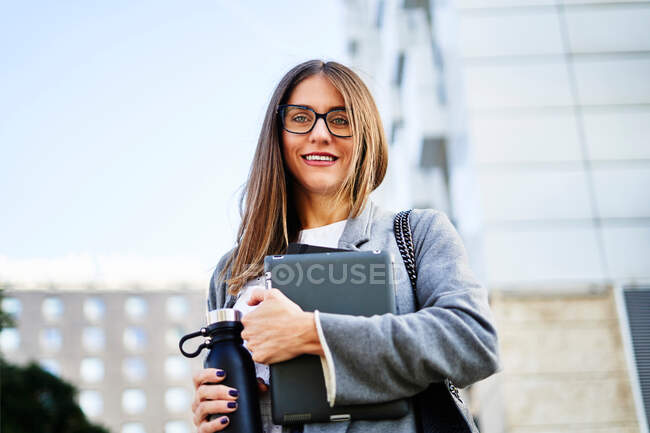 Снизу положительная женщина-предприниматель в каштановых волосах в стильном наряде смотрит в камеру, стоя с планшетом и термосом на городской улице против здания в дневное время — стоковое фото