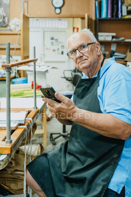 Konzentrierte Seniorchefin in Schürze und Brille mit Handy während des Druckvorgangs im Atelier an der Werkbank sitzend — Stockfoto