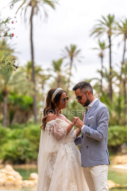 Seitenansicht von glücklichen jungen Eheleuten in Hochzeitskleidern stehend und Händchenhaltend, während sie sich am See und bei Tag im Garten mit grünen Bäumen betrachten — Stockfoto