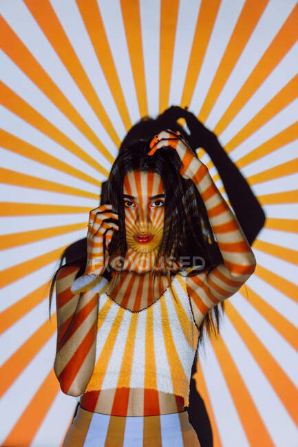 Fresco giovane donna etnica in crop top con strisce sul corpo dalla luce del proiettore guardando la fotocamera — Foto stock