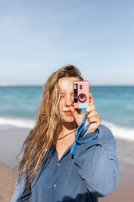 Young female in wet clothes taking photo on camera while standing looking at camera on sandy beach near waving sea — Stock Photo