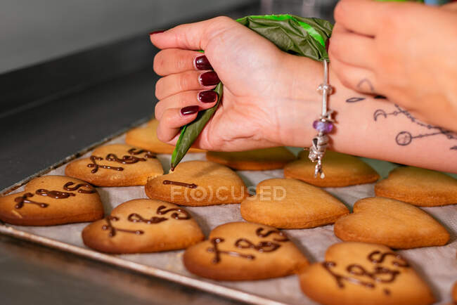 Crop young female confectioner decorating heart shaped sugar cookies with chocolate icing standing at table in bakery — Stock Photo