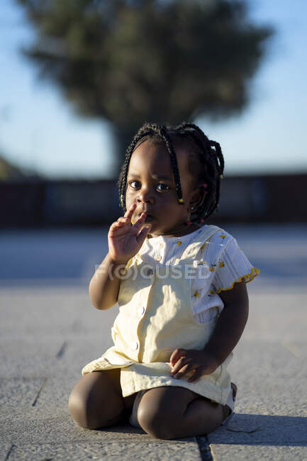 Calma niña afroamericana con trenzas negras en traje de moda lamiendo dedo y mirando hacia otro lado mientras está sentado en la pasarela de asfalto en la calle en el día soleado - foto de stock