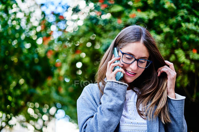 Trabajadora positiva con el pelo rubio de pie en la acera cerca de los coches y hablando por teléfono celular mientras mira hacia abajo con una sonrisa dentada - foto de stock