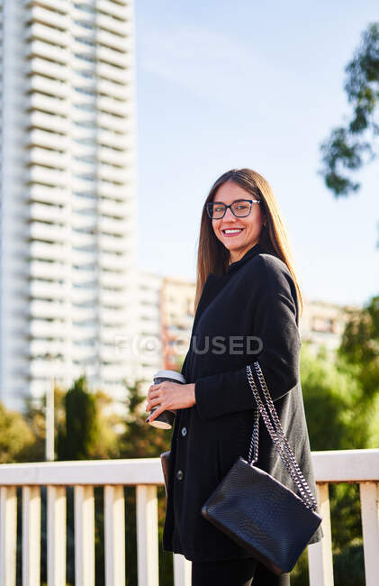 Снизу положительная молодая предпринимательница в черном пальто, стоящая с мешком и чашкой горячего напитка на вынос и широко улыбающаяся — стоковое фото
