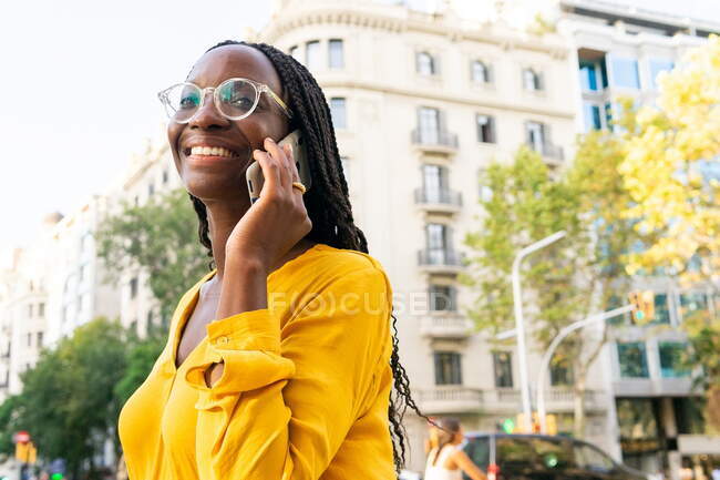 Mulher afro-americana positiva em óculos olhando para longe ao ter uma conversa telefônica na rua com edifícios residenciais e árvores na cidade — Fotografia de Stock