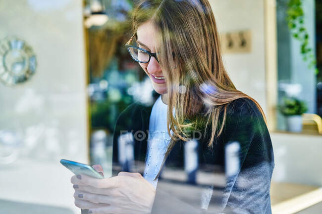Grâce à un verre de culture employé féminin positif dans les lunettes à l'aide d'un téléphone mobile pour vérifier les messages de travail — Photo de stock