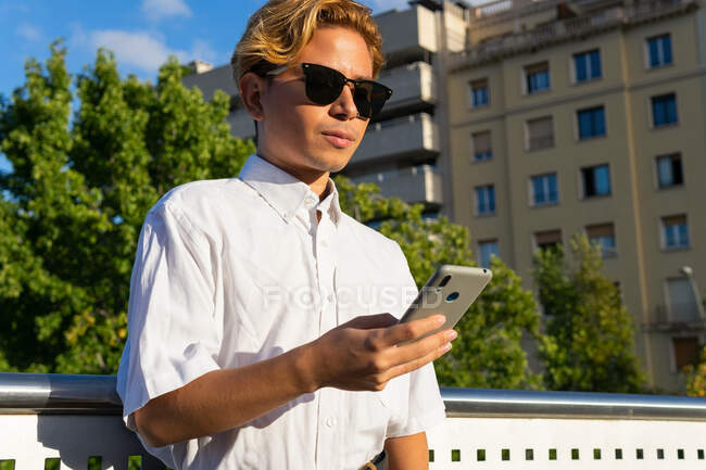 Selbstbewusster junger Mann in weißem Hemd, der an einem sonnigen Tag auf der Straße vor blauem Himmel steht und SMS aufs Handy sendet — Stockfoto