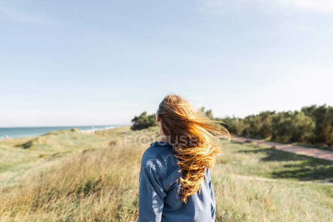 Обратный вид на неузнаваемую молодую женщину в повседневной одежде, стоящую на травянистом лугу летом и смотрящую в камеру — стоковое фото