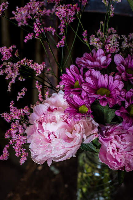 Bouquet de pivoines colorées fraîches et de chrysanthèmes dans un vase en verre placé sur une chaise en bois altérée près de rideaux dans une pièce lumineuse — Photo de stock