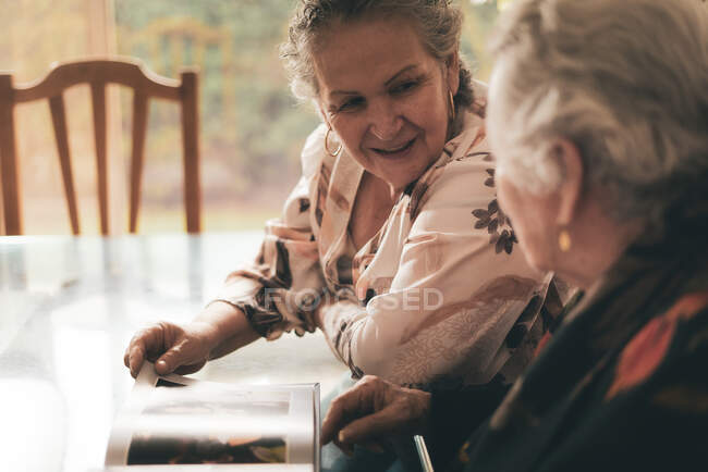 Sœurs âgées inspectant des photos dans un album photo et discutant de souvenirs assis à table à la maison ensemble — Photo de stock