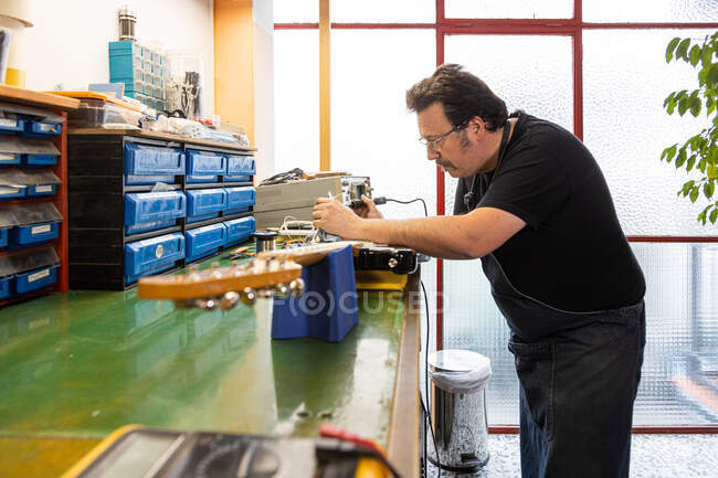 Seitenansicht eines konzentrierten männlichen Meisters mit Brille und Arbeitskleidung, der E-Gitarre repariert, während er am Tisch in der professionellen Werkstatt steht — Stockfoto