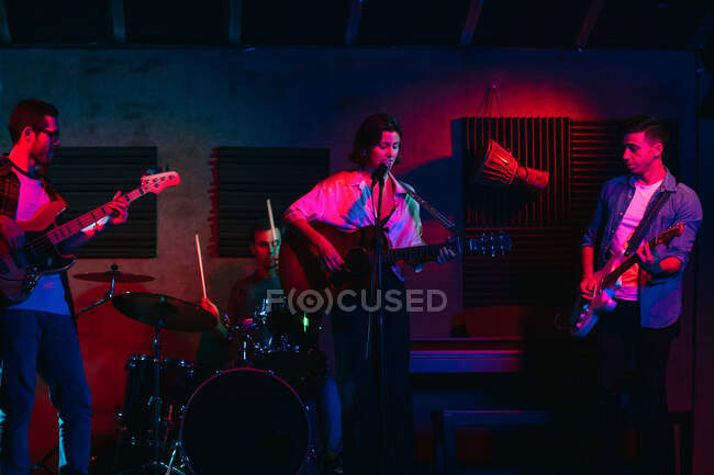 Grupo de personas en ropa casual tocando guitarras y tambores mientras mujer cantando y cantando canción en el club con luces de neón - foto de stock