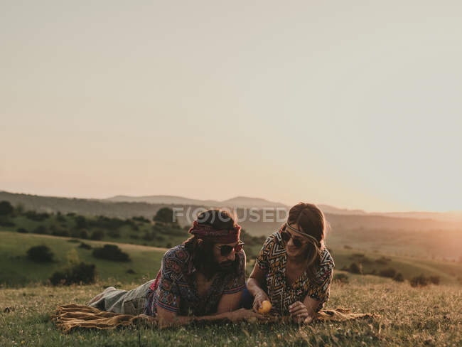 Пара хіпі з сонцезахисними окулярами, що взаємодіють, лежачи на трав'янистих лугах в природі під час заходу сонця — стокове фото