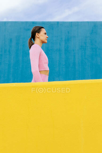 Vue latérale de la femelle concentrée sérieuse aux cheveux foncés portant des vêtements de sport rose clair regardant loin contre le mur bleu — Photo de stock