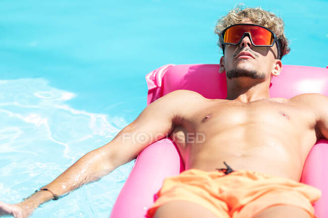 Hombre tranquilo en pantalones cortos de natación y gafas de sol tomando el sol en el colchón inflable rosa en la piscina con agua clara en el día soleado del verano - foto de stock