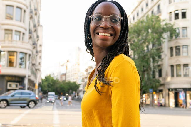 Позитивная афроамериканка в очках с черными волосами стоит на улице с жилыми домами в городе против безоблачного неба — стоковое фото