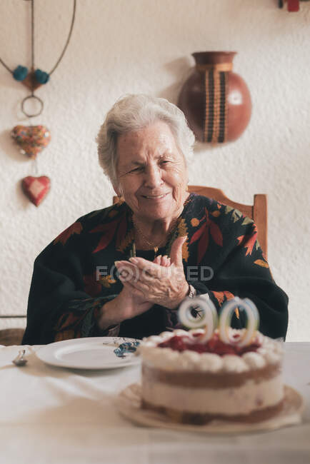 Aged woman blowing candles on birthday cake then clapping hands while celebrating 90th anniversary with relative at home — Stock Photo