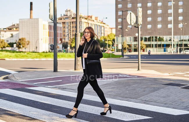 Full length of cheerful female entrepreneur in formal clothes talking on smartphone while crossing road on city street in sunny day — Stock Photo