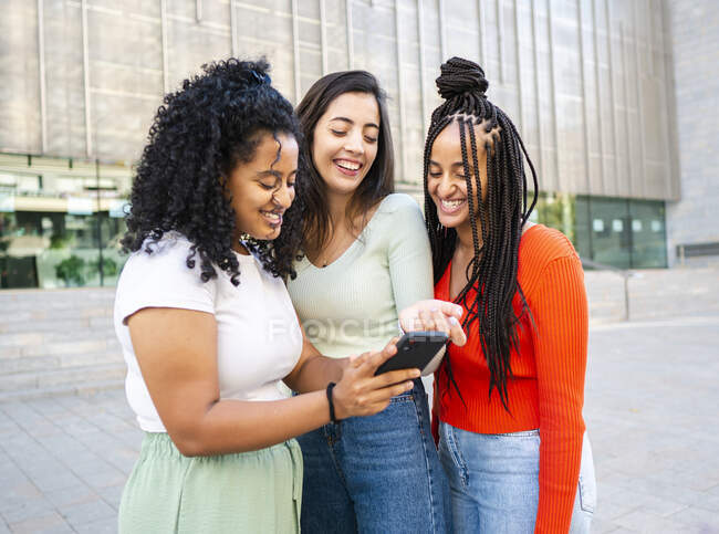 Cheerful multiracial girlfriends in trendy clothes laughing and showing video on smartphones on city street in daylight — Stock Photo