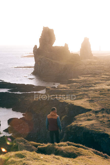 Vista posteriore di anonimo escursione maschile in caldo outerwear in piedi su una scogliera rocciosa e guardando lontano contro Reynisdrangar basalto steli del mare al tramonto in Islanda — Foto stock