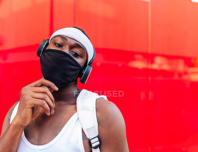 Homme afro-américain sérieux en masque de protection et écouteurs sans fil debout contre le mur rouge dans la rue de la ville pendant la pandémie COVID et regardant la caméra — Photo de stock