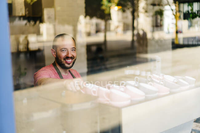 Through glass of male worker in apron taking cups from shelf while standing near window during work in modern cafeteria — Stock Photo