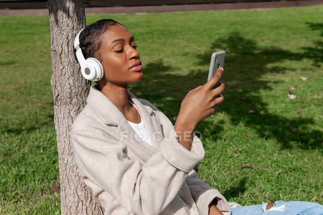 Tranquila mujer afroamericana con los ojos cerrados escuchando música en auriculares inalámbricos mientras está sentada en el césped cerca del tronco del árbol en el soleado parque mientras usa el teléfono inteligente - foto de stock