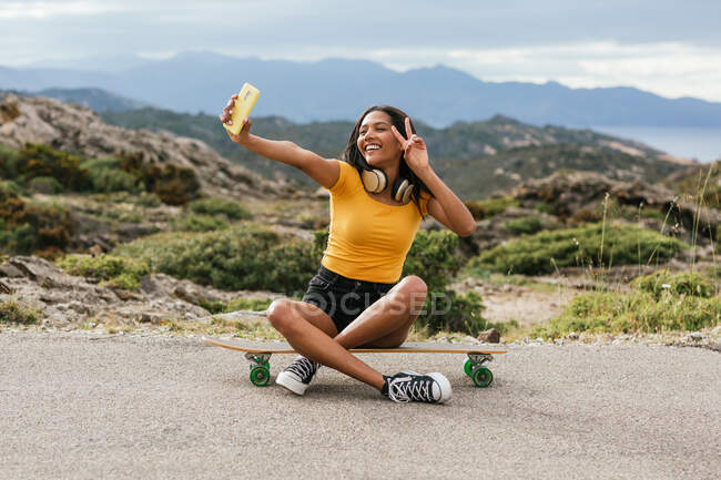 Full body of happy ethnic female sitting on longboard with crossed legs while showing peace gesture and taking self portrait on cellphone — Stock Photo