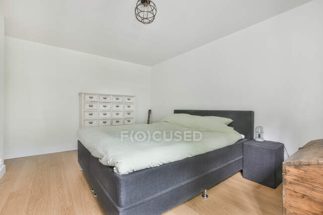 Bett mit weißer Bettwäsche an der Wand mit Nachtlicht auf dem Nachttisch im hellen Schlafzimmer mit Schubladen in der Wohnung — Stockfoto