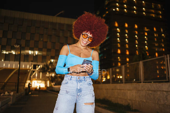 Позитивна жінка з зачіскою в африканському стилі, одягнута в модний одяг, надсилає повідомлення на мобільний телефон, стоячи на вулиці з будинками в нічний час. — стокове фото
