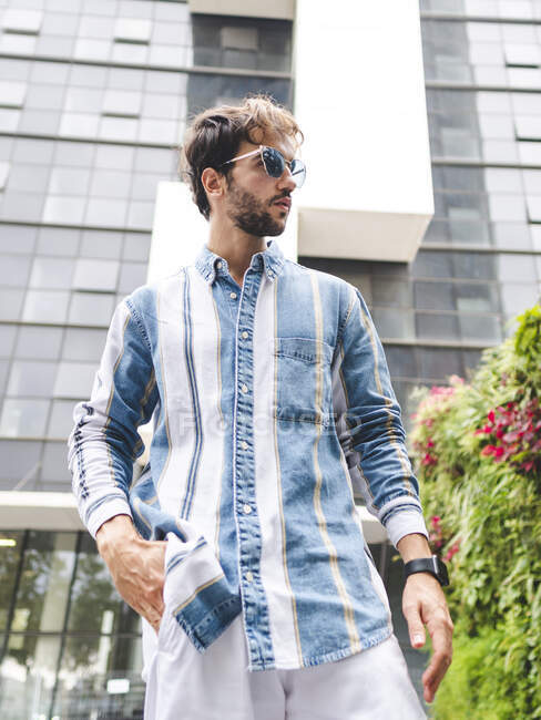 Low angle of young male in sunglasses standing with hand in pockets and looking away on street against building — Stock Photo