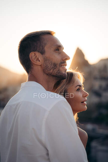 Vista laterale di abbracciare l'uomo e la donna innamorati in piedi insieme sulla strada e guardando lontano contro il tramonto — Foto stock