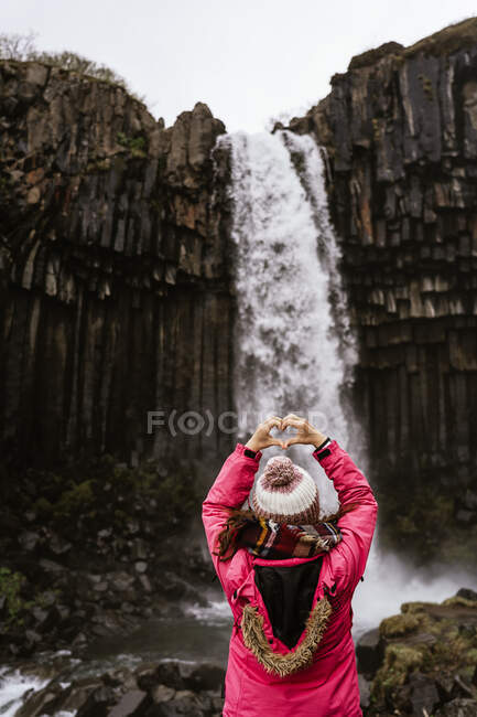 Rückansicht einer unkenntlichen Touristin in warmer Kleidung und Hut, die eine Herzensgeste zeigt, während sie in der Nähe des mächtigen Svartifoss-Wasserfalls steht, der durch Basaltklippen fließt und die Natur bewundert — Stockfoto