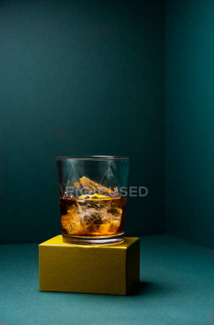 Front view of a glass cup with cold whiskey and cubes of ice placed on a gold-colored rugged base and a turquoise blue corner background — Stock Photo