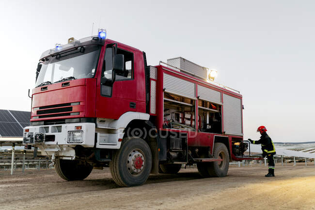 Modern red fire rescue truck equipped with fire extinguisher and hoses with bright flashlight on top and anonymous firefighter wearing uniform standing pulling out a portable light generator — Stock Photo