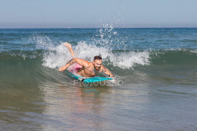 Hombre activo en camisas de baño acostado en la tabla de surf mientras practica surf en el mar ondulante en el resort tropical en el soleado día de verano - foto de stock
