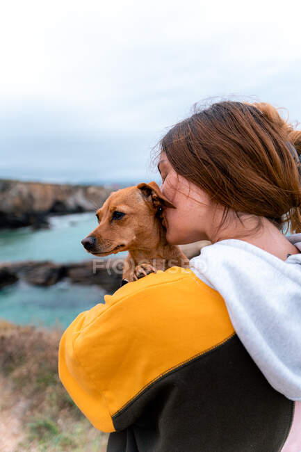 Feliz mujer viajera besando lindo cachorro mientras está de pie en el prado en las tierras altas en la playa de las catedrales en Galicia al atardecer - foto de stock