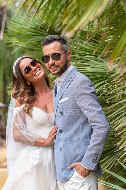 Усміхнена молода подружня пара у весільному одязі та сонцезахисних окулярах стоїть біля зелених дерев та рослин в літній день у парку — стокове фото
