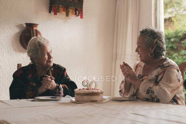 Mujer anciana con cabello gris y mujer mayor sentada en la mesa de comedor y celebrando 90 cumpleaños con delicioso pastel con velas - foto de stock