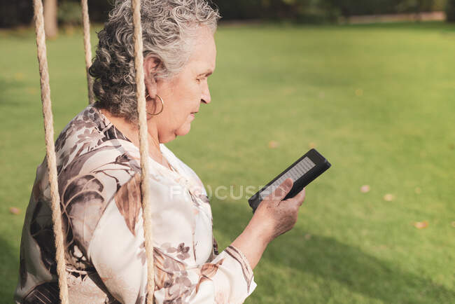 Vista lateral de la señora mayor con blusa sentada en el parque y leyendo libro electrónico - foto de stock