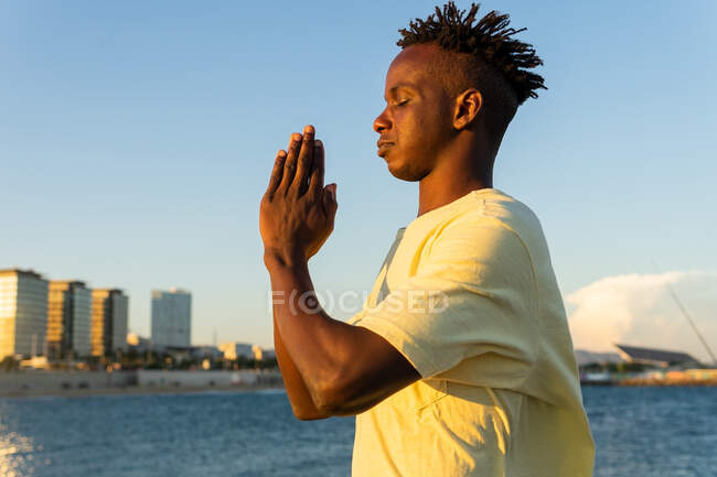 Vista lateral do pacífico afro-americano masculino em roupas casuais de pé com os olhos fechados e fazendo gesto namaste com as mãos na rua no verão — Fotografia de Stock