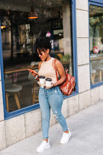 Повний набір трендів молодих жінок тисячоліття з темним африканським волоссям в стильному одязі і рюкзаком з використанням мобільного телефону під час ходьби по міській вулиці з чашками кави. — стокове фото