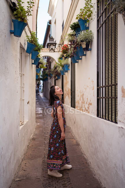 Vista laterale del corpo pieno di sorridente turista asiatica in piedi in stretto passaggio tra le case mentre osserva vasi da fiori in città — Foto stock