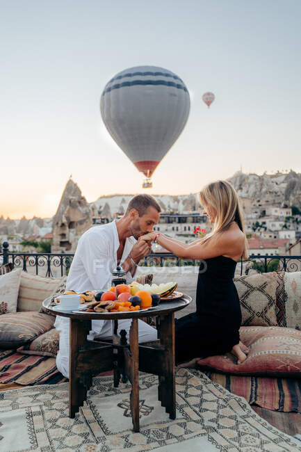 Mann küsst Hand seiner Freundin, während er auf dem Dach der Stadt sitzt und Heißluftballons in den Abendhimmel steigen — Stockfoto