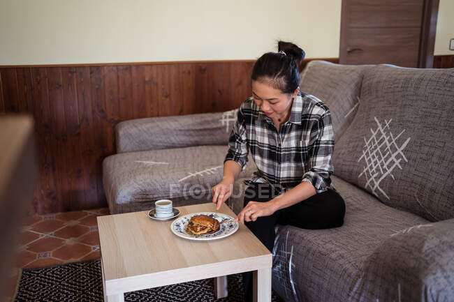 Jovem etnia asiática fêmea comendo panquecas caseiras colocadas na placa perto da xícara de café na mesa enquanto se senta no sofá confortável na sala de estar — Fotografia de Stock