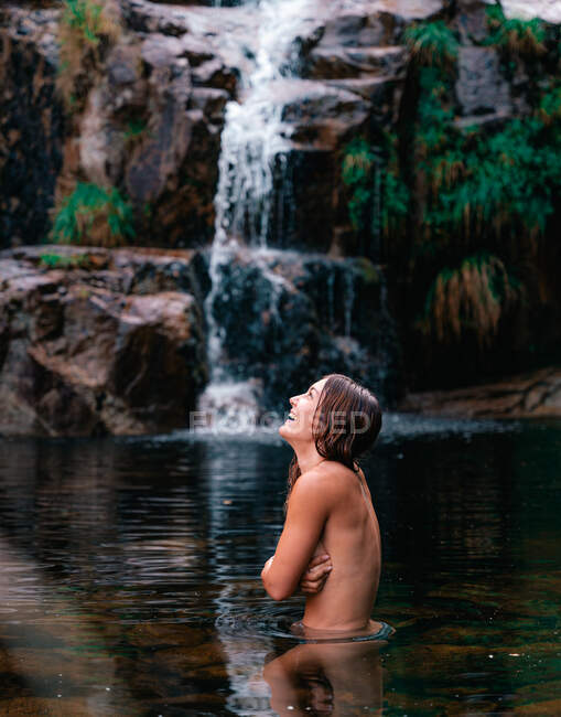 Vista laterale di sorridere viaggiando nudo seno femminile di copertura e in piedi in acque calme di stagno vicino alla cascata nel parco naturale di Fervenza De Casarinos in Spagna — Foto stock