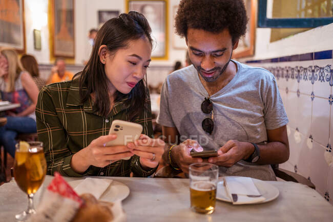 Jovem casal multirracial feliz em roupas casuais usando smartphones enquanto se sentam juntos à mesa com copos de cerveja no restaurante moderno — Fotografia de Stock