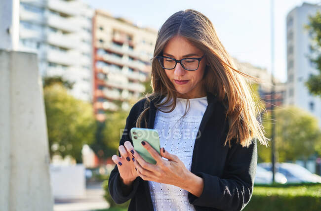 Знизу зосереджена бізнес-леді в модному одязі і окулярах стоїть і переглядає смартфон проти зелених дерев в парку в літній час в денний час — стокове фото