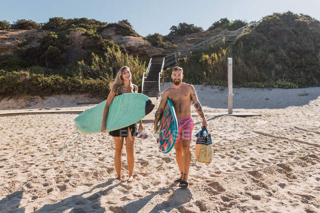 Corpo inteiro de casal esportivo feliz com pranchas de surf passeando juntas na praia de areia antes de treinar no mar — Fotografia de Stock